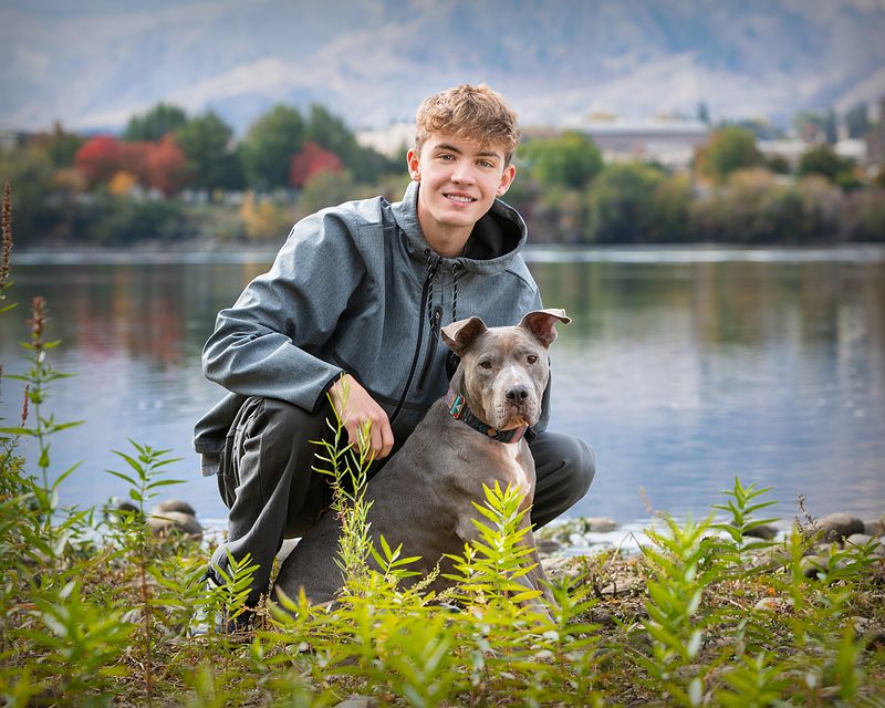 Senior crouching beside a dog on the riverbank, with autumn trees behind